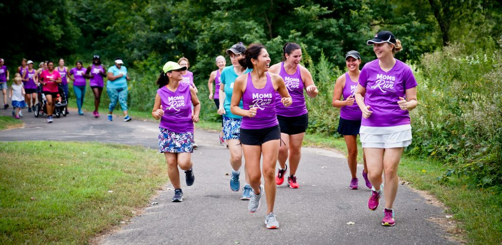 A group of Moms on the Run wearing matching purple shirts run on a path in the woods.